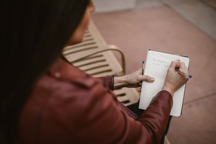 A woman in a brown leather jacket writing in a notebook, captured outdoors.