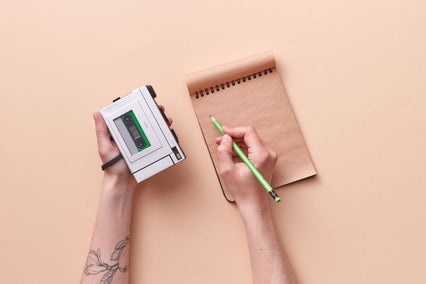 Vintage cassette player held in hand with notepad and pencil, on a minimalist pastel background.